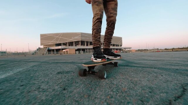 Young skateboarder balances on board in spacious urban plaza near modern stadium, seen from low angle with tattooed arms, streetwear, camouflage pants, chunky sneakers, pastel dusk sky, and empty