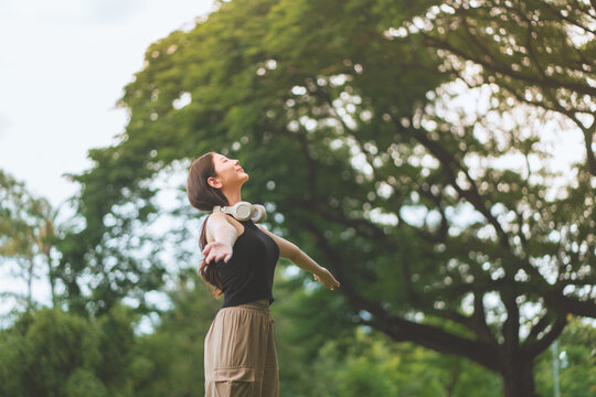 Young woman with headphones around neck spreading arms and relaxing in green nature, enjoying freedom, mindfulness, fresh air, peaceful outdoor wellness, positive energy and healthy lifestyle