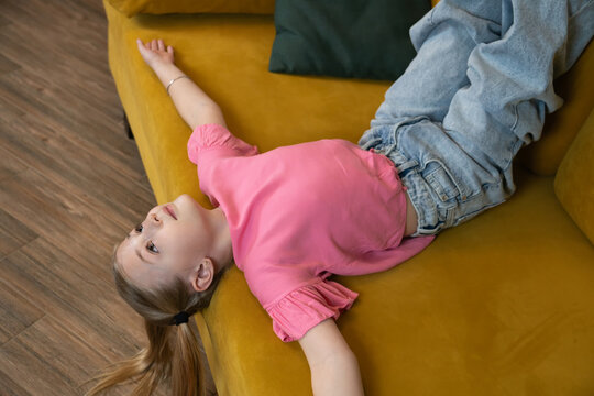 Young child girl lying relaxed on a vibrant yellow sofa, looking upwards with a thoughtful expression, depicting concepts of childhood innocence, contemplation, and peaceful rest