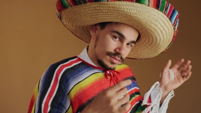 Young man wearing large woven straw sombrero with bright striped trim and matching multicolor serape-style poncho reaches toward camera with open hand and relaxed wrist while smiling directly at