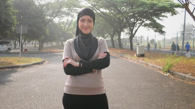 Confident young Asian Muslim woman stands with arms crossed on a tree-lined road in a park, smiling and projecting strength, modest activewear and a healthy outdoor lifestyle.