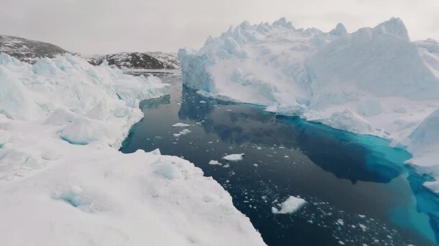Dynamic aerial drone flight navigating low over calm blue water, passing close to massive icebergs and floating ice chunks in a polar landscape