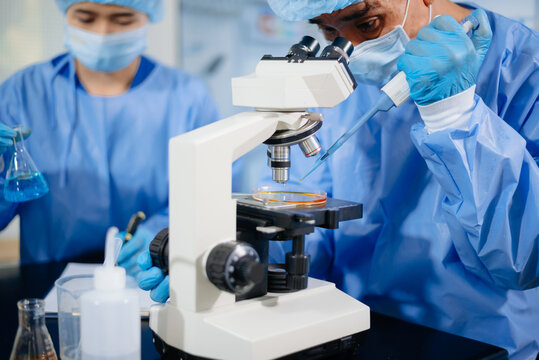 Two dedicated medical scientists in surgical protective gowns using a micropipette and microscope in research laboratory.