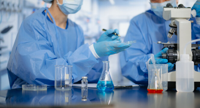 Two dedicated medical scientists in surgical protective gowns using a micropipette and microscope in research laboratory.