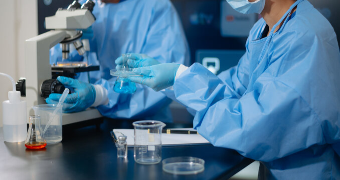 Two dedicated medical scientists in surgical protective gowns using a micropipette and microscope in research laboratory.