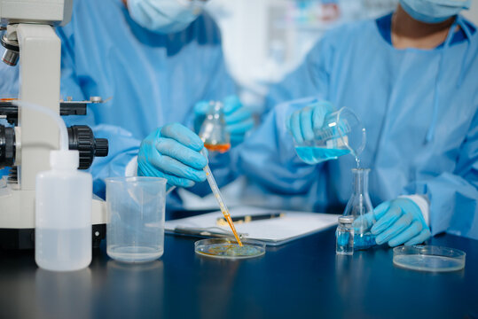 Close up of medical researchers in blue protective gowns using a pipette and beaker scientific testing near a microscope in a laboratory.