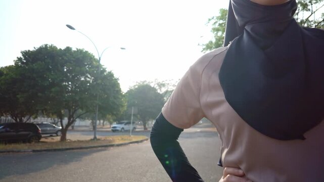 Young Asian Muslim woman wearing a black hijab and sportswear stands outdoors at sunrise with hands on hips, conveying confidence, fitness, modest fashion and an active, healthy lifestyle.
