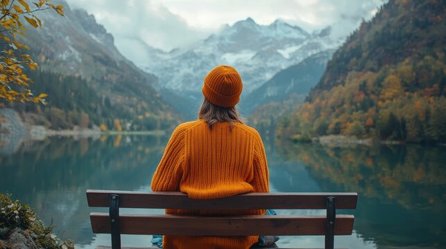 Profile of a woman wearing an orange sweater and cap seated on a wooden bench, appreciating the stunning landscape of a mountain range and tranquil lake