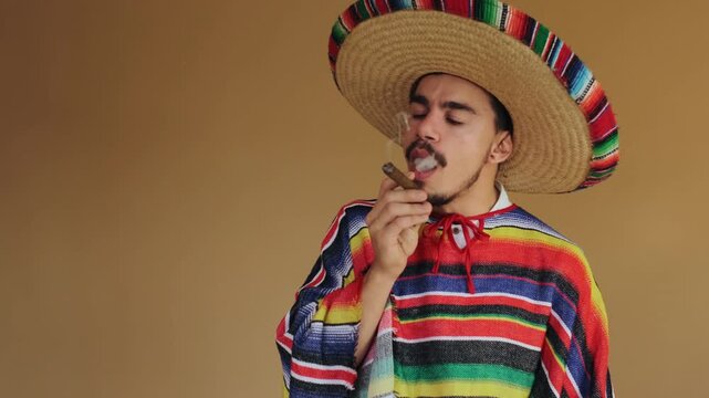 Young man wearing wide woven straw sombrero with multicolor brim and brightly striped serape poncho holds lit cigar near chest while looking toward camera and exhaling thin curl of smoke