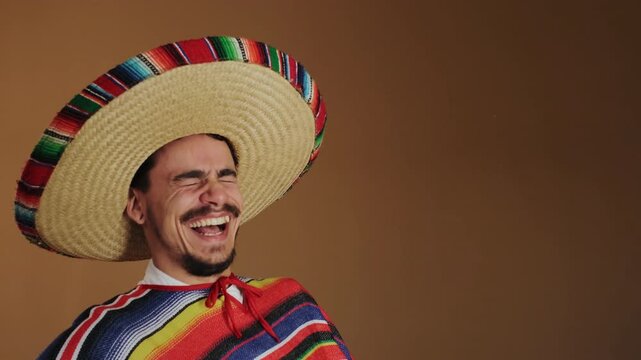 Close view of smiling man wearing oversized braided straw sombrero and colorful striped poncho, laughing with bright eyes, visible teeth, mustache, goatee, against warm brown studio background