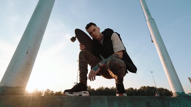 Low-angle view shows young male skateboarder crouched on concrete ledge, holding board close beside body between tall poles, with skateboarder, young, male, crouching detail, showing board, concrete