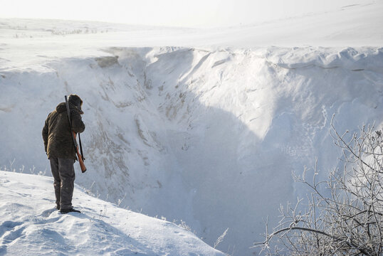 A hunter with a shotgun stands on the very edge of a deep snow-covered ravine on a winter day.