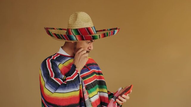 Adult male wearing wide-brimmed woven straw sombrero with multicolored band and matching striped serape poncho taps orange smartphone with left index finger while holding device in right hand