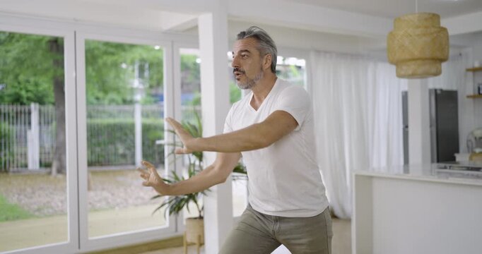 Senior caucasian middle aged man graceful martial arts movements in a bright modern home, promoting balance wellness health care, mindfulness fitness, and healthy living work out longevity activity