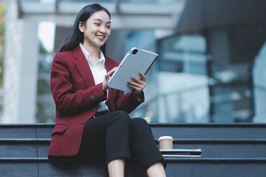 Focused woman in blazer types on laptop outdoors. Urban professional works remotely on stairs. She uses modern tech for business and connection.