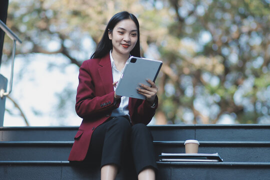 Focused woman in blazer types on laptop outdoors. Urban professional works remotely on stairs. She uses modern tech for business and connection.