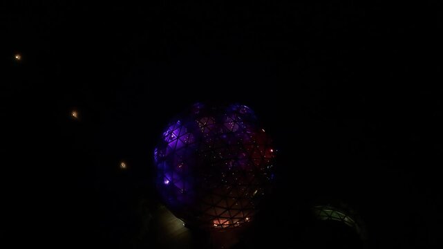 Stunning aerial drone video showcasing a glowing geodesic dome lit with vivid purple and orange lights against a dark nightscape, featuring modern architecture and dramatic contrasts.