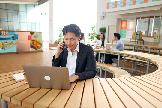 Asian Businessman Talking on Smartphone While Working on Laptop in Modern Courtyard