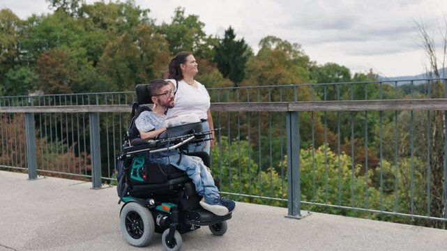 Powered wheelchair user and Personal assistant moving together along an outdoor walkway, enjoying accessible mobility surrounded by nature.
