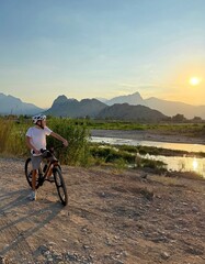 A boy wearing a helmet stands with his mountain bike on a rocky dirt path, looking out over a river valley toward distant mountains at sunset. The scene captures the spirit of active outdoor adventure © happy_artist