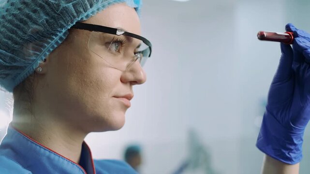 Female medical worker in side profile studies blood sample tube at eye level inside blurred laboratory, with blue scrubs, hair cover, safety glasses, and gloves, creating precise clinical mood and
