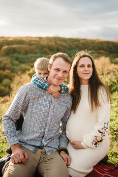 Family portrait outdoors in autumn. A pregnant mother, father, and their little son. The son is hugging his dad from behind, with mountains in the background. Closeup.