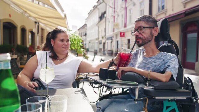 Personal assistant helping a wheelchair user enjoy a refreshing drink while relaxing at an outdoor cafe. Disability and community inclusion.