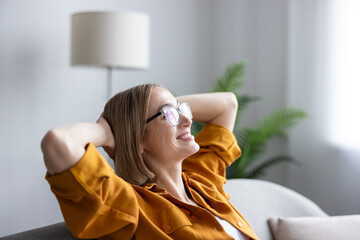 Obraz premium Young woman with eyeglasses relaxing on a sofa with hands behind her head, enjoying a moment of comfort and quiet happiness after achieving success at her remote work