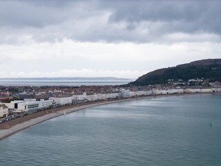 Fototapeta premium Llandudno aerial photo in summer looking across the curved North Wales seafront with promenade coastal buildings calm bay Great Orme hillside and dramatic cloudy sky above