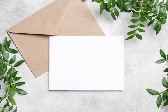 Blank white card with a brown kraft envelope on a light concrete surface, surrounded by fresh green leaves. Ideal for invitation and greeting card mockups