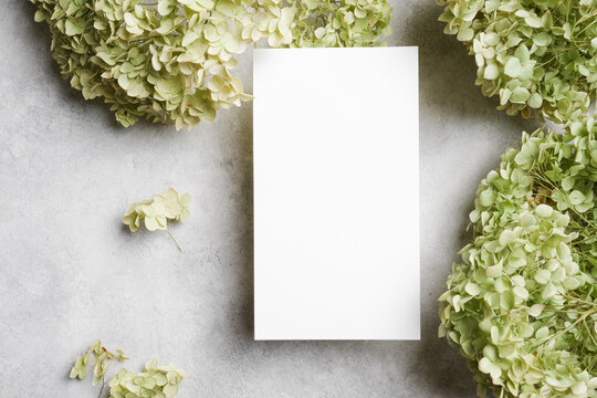 Blank vertical menu card mockup on gray stone surface, surrounded by soft green hydrangeas, ideal for elegant dining or wedding stationery