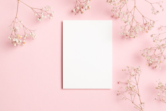 Vertical white blank card mockup framed by soft white baby breath flowers on pastel pink background, perfect for wedding invitations or cards, flat lay