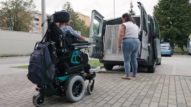 Personal assistant deploying a wheelchair ramp and assisting a man with disability entering an adapted vehicle.