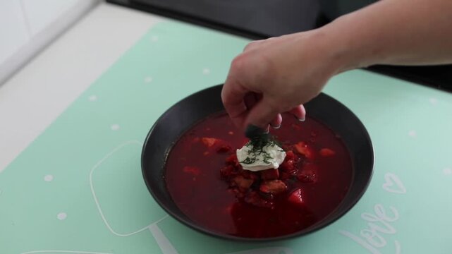 Woman serving traditional hot borscht from a cooking pot into a bowl in a home kitchen. 