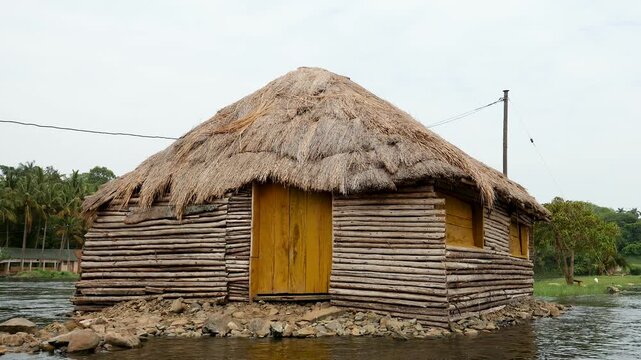 Unique traditional wooden hut with a thatched grass roof standing on a small rocky island in the middle of a tranquil river, showcasing rural african architecture and a serene natural landscape