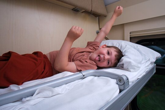 Happy child lying on upper bunk in train compartment. Little boy having fun during railway travel. Excited kid showing tongue while resting on bed in passenger carriage during trip.