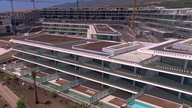 Aerial view of a large residential construction site with modern apartment buildings, rooftop terraces, swimming pools and cranes showing ongoing development progress