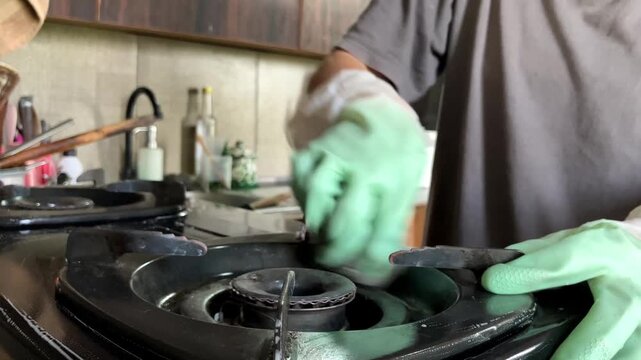 Slow motion close up video with selective focus on a black gas stove. A housewife wearing rubber gloves is wiping and cleaning the dirty kitchen appliance for daily domestic chores and hygiene.