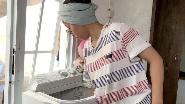 Slow motion video of an Asian housewife wearing a headband and glasses. She is busy doing her daily domestic chores by washing clothes in a top load washing machine at home.