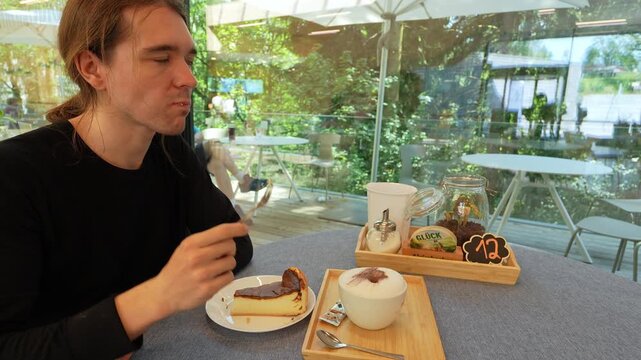 A man savors delicious cheesecake and coffee inside a bright cafe in Germany. Glass walls offer a tranquil view of lush green trees and an outdoor area, perfect for a relaxing break.