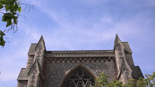 Static low-angle view of the Shrine of St Alban east gable with flint stonework, pinnacles, and tracery window beneath a light sky.