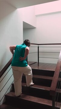 senior asian female slowly climbing staircase with back pain, indoor home scene during daytime, health, A11Y, and mobility concept, indoor scene with one 60s old woman, back view,  vertical video