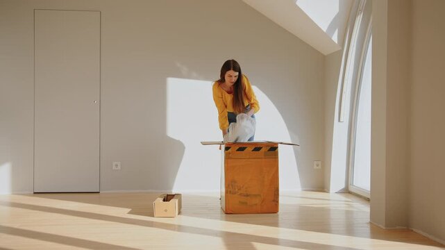 Woman in yellow sweater and blue jeans carefully packs items into cardboard boxes in a bright, sunlit room