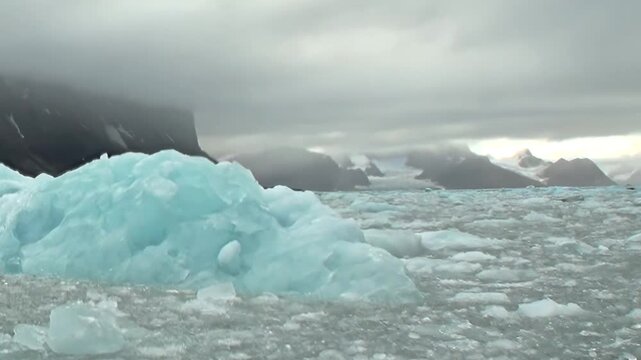 Embark on extreme travel experience across Arctic Ocean. Brilliant blue icebergs and countless ice floes create mesmerizing, frozen path. Dark, imposing mountains rise beneath cloudy, ethereal sky.