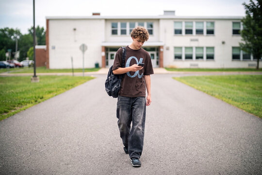 A teenager at school. A teenager with a backpack walks along the road from the school building, carefully looking at the smartphone screen. High school student, internet, gadgets and modern lifestyle