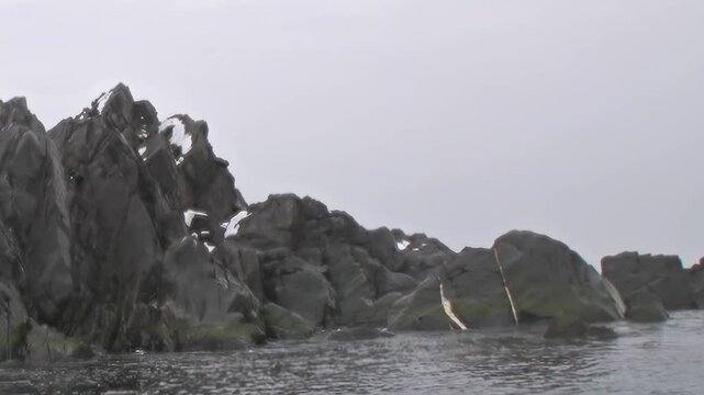 Jagged, snow-dusted rock formations majestically pierce the dark, frigid Arctic Ocean waters. Grey skies loom above this isolated wilderness, marking a journey through nature's raw, extreme frontier.