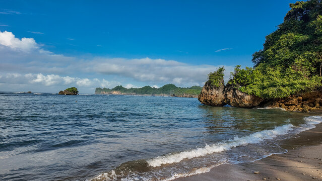 Under the clear blue sky, admire the stunning tropical beach filled with sand, rocks, coconut trees, and tourists. Kondang Merak Beach, Malang, Indonesia.