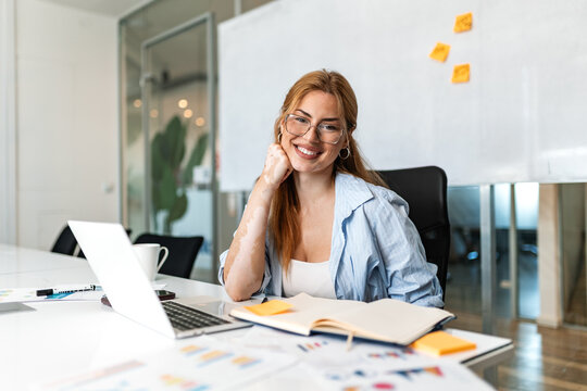 Happy businesswoman at office desk. Smiling female entrepreneur looking at camera with hand on chin next to laptop, notebook and charts in modern workplace. Business success concept.