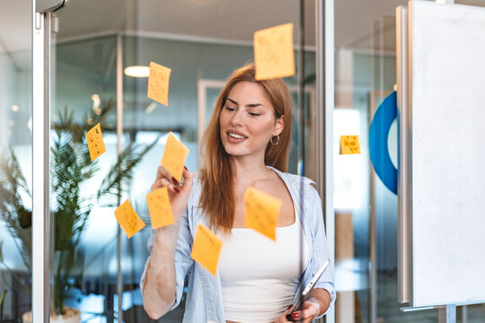 Smiling businesswoman organizing sticky notes on glass wall. Female professional planning project with adhesive memos and tablet in modern office. Strategy concept.