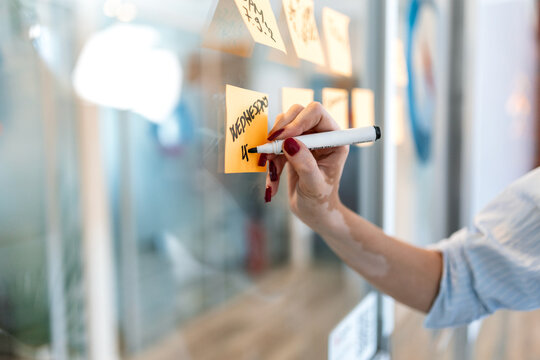 Businesswoman writing on sticky note on glass wall. Focused female professional planning project with adhesive notes in modern office. Strategy and organization concept.